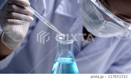 Professional scientist wear protective glass and medical gloves to analyze blue essential, which dropped into an erlenmeyer flask from plastic pipette in medical laboratory. Front view, copy space Professional scientist wear protective glass and medical gloves to analyze blue essential, which dropped into an erlenmeyer flask from plastic pipette in medical laboratory. Front view, copy space 117651552