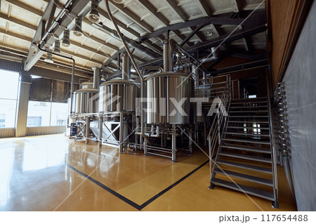 Large, modern brewery with several stainless steel tanks, connected by a network of pipes and valves, with staircase leading to platform above 117654488
