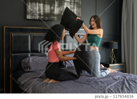 Playful young black and white female couple having pillow fight on bed in bedroom at home in the morning 117654816