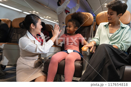 Airplane flight attendant providing service to kid and mother passengers on board. Asian female air hostess giving high five to African kid girl on board. 117654818