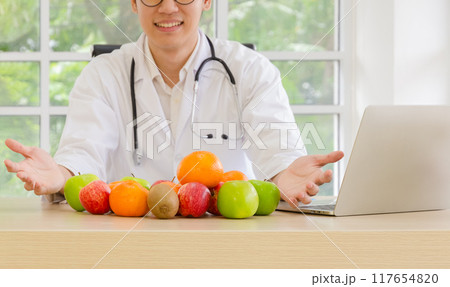 Male doctor in white coat with stethoscope at office desk with laptop and presenting variety of fresh fruits including apples, oranges and kiwis. 117654820