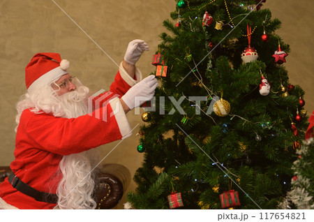 Santa Claus sitting on leather sofa and reaching up to hang ornaments on decorates Christmas tree at home. 117654821