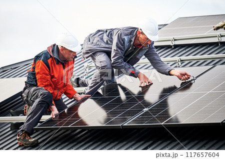 Roofers installing photovoltaic solar panels on roof of house. Men engineers in helmets building solar module system with help of hex key. Concept of alternative, renewable energy. Roofers installing photovoltaic solar panels on roof of house. Men engineers in helmets building solar module system with help of hex key. Concept of alternative, renewable energy. 117657604