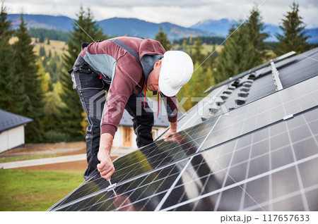 Man installer mounting photovoltaic solar panels on roof of house. Engineer in helmet installing solar module system with help of hex key. Concept of alternative, renewable energy. 117657633