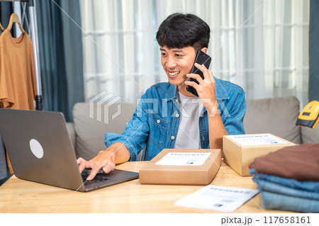 Happy young online seller man talking to his customer on the phone while checking incoming orders from his laptop computer 117658161