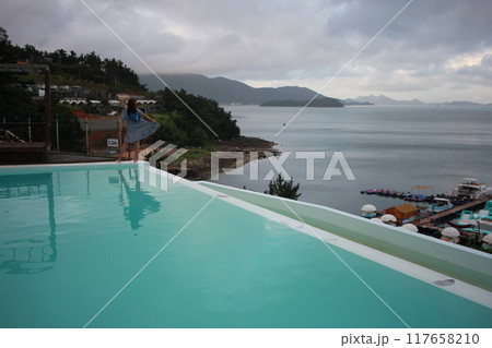 Dolsan-eup, Yeosu-si, South Korea - July 26, 2024: Young woman watching sunrise at a resort with a swimming pool overlooking the sea in Yeosu Dolsan-eup, Yeosu-si, South Korea - July 26, 2024: Young woman watching sunrise at a resort with a swimming pool overlooking the sea in Yeosu 117658210