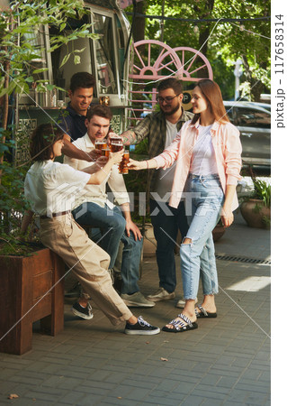 Warm sunny day and relaxing vibes with friends, Happy, smiling young people, men and women meeting at outdoor bar, restaurant and enjoying beer 117658314