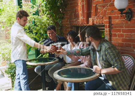 Lively group of young people, men and women, friends gathering at local outdoor bar, clinking beer glasses, enjoying good company Lively group of young people, men and women, friends gathering at local outdoor bar, clinking beer glasses, enjoying good company 117658341