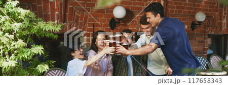 Smiling happy young people, men and women, friends gathering at outdoor bar in warm sunny evening and enjoying refreshing beer. Cheers. 117658343