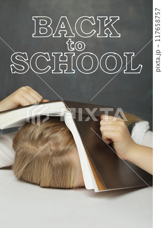 Little child girl pupil hiding under textbook in classroom against white Back to school inscription 117658757