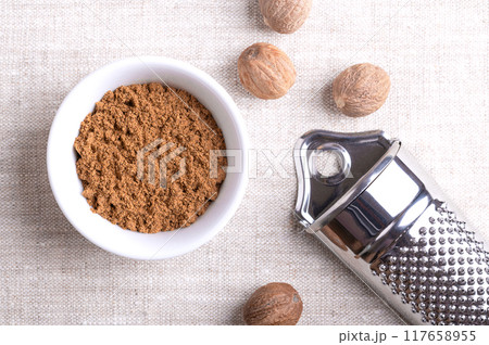 Nutmeg powder, ground nutmeg in a white bowl on linen fabric. Fragrant or true nutmeg, a spice, also used in traditional medicine. Beside the bowl dried seeds of Myristica fragrans and nutmeg grater. Nutmeg powder, ground nutmeg in a white bowl on linen fabric. Fragrant or true nutmeg, a spice, also used in traditional medicine. Beside the bowl dried seeds of Myristica fragrans and nutmeg grater. 117658955