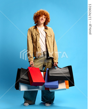 Young man, student dressed stylishly holding heavy shopping bags against blue studio background. Mega discount. Concept of Black Friday sale. 117659021