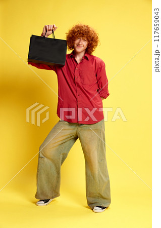 Delightful young man dressed vintage stylish outfit holding black back with purchases against yellow background. Concept of Black Friday sale. 117659043
