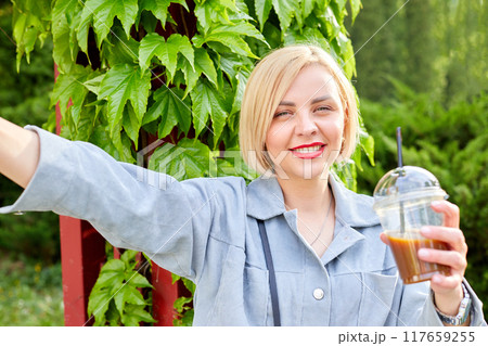 Stylish young blonde walks around the city, drinks a cold drink and takes a selfie on her mobile phone. 117659255