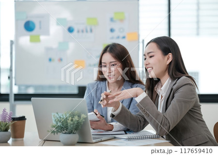 Two women are sitting at a table with a laptop and a whiteboard 117659410