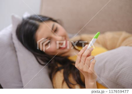 A woman is laying on a couch with a thermometer in her hand A woman is laying on a couch with a thermometer in her hand 117659477