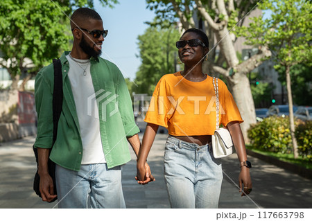 Joyful african american loving couple walking hold hands in city street guy makes girlfriend laugh. 117663798