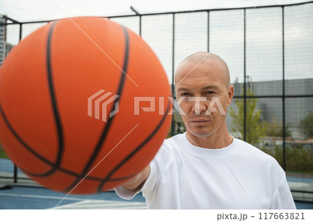 Portrait of sporty adult man holding orange basketball ball in hand on sports court. Focus on man. Close up. 117663821