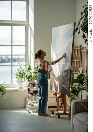 Peaceful woman artist in rays of bright light warm sun moving easel with large canvas to side. Peaceful woman artist in rays of bright light warm sun moving easel with large canvas to side. 117663922