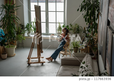 Creative happy female artist in home art studio filled with green flowers and plants in pots. 117663936
