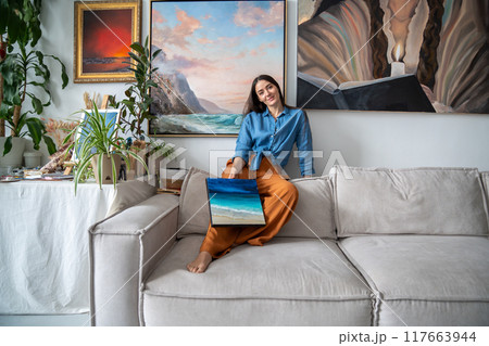 Creative satisfied female artist demonstrates picture, relaxed sitting on couch at home art studio. 117663944