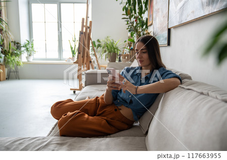 Peaceful pleased woman using smartphone, procrastinating relaxing resting on couch at home workshop 117663955