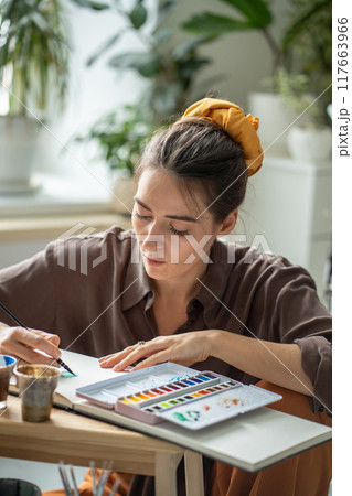 Creative woman artist painting on paper of sketchbook with watercolour sits on floor in art studio. 117663966