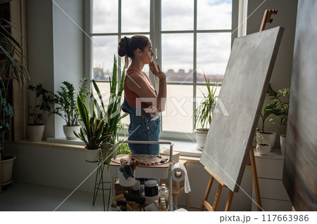 Serious pensive artistic female in front of window in art studio reflecting before starting work. Serious pensive artistic female in front of window in art studio reflecting before starting work. 117663986
