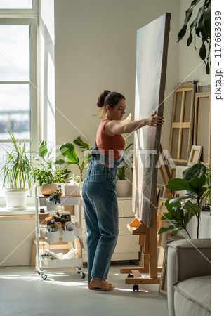 Peaceful woman artist in rays of bright light warm sun moving easel with large canvas to side. 117663989