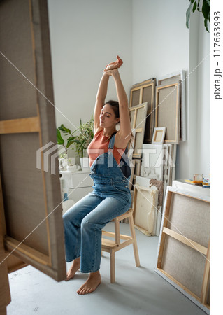 Tired artist girl stretching hands relaxed from painting on canvas on easel in art craft studio. 117663993