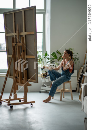 Dreaming creative woman artist take coffee break in art studio eat sandwich looks on canvas on easel 117664060