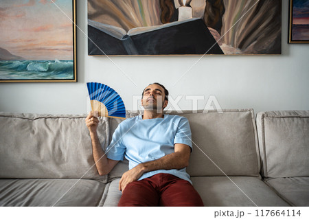 Exhausted unhappy man blowing with paper fan, suffering from stuffiness heat, lying on sofa. Exhausted unhappy man blowing with paper fan, suffering from stuffiness heat, lying on sofa. 117664114