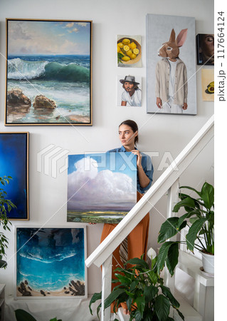 Serious pensive woman with canvas makes creative art composition from paintings on studio wall. 117664124