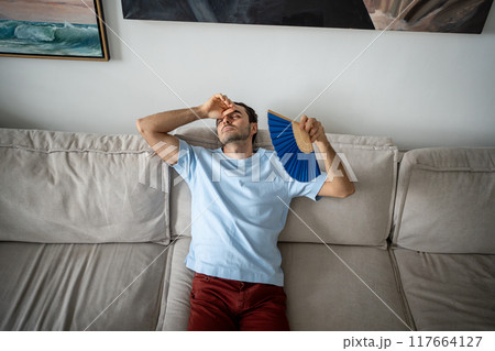 Exhausted unhappy man blowing with paper fan, suffering from stuffiness heat, lying on sofa. Exhausted unhappy man blowing with paper fan, suffering from stuffiness heat, lying on sofa. 117664127