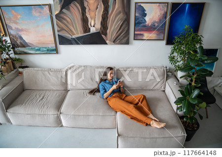 Slothful girl procrastinating lying on sofa with smartphone. Woman relaxing at home art studio. 117664148