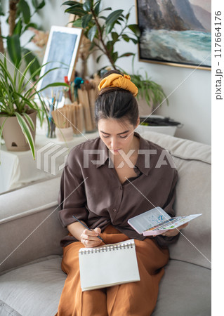 Inspired artistic woman calmly carefully drawing sketch with paintbrush, watercolors paints. 117664176