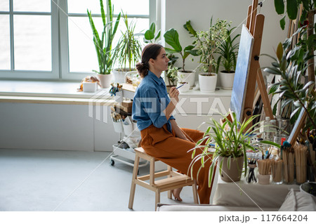Unsmiling female artist working on creative painting sitting in light white art home studio. Unsmiling female artist working on creative painting sitting in light white art home studio. 117664204