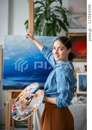 Satisfied pleased artistic woman working on painting, drawing details with small paintbrush. Satisfied pleased artistic woman working on painting, drawing details with small paintbrush. 117664206