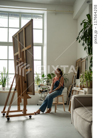 Joyful woman artist looking at huge canvas admiring enjoying examining sitting at home art studio. Joyful woman artist looking at huge canvas admiring enjoying examining sitting at home art studio. 117664208