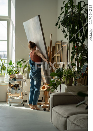 Interested artist young woman holds canvas puts on easel to continue painting in oil in art studio. 117664212