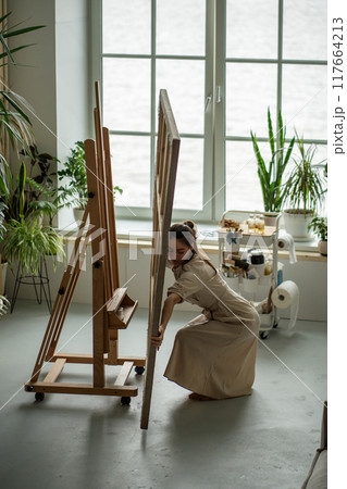 Female artist in bright home art studio organizing preparing workplace, putting workspace in order Female artist in bright home art studio organizing preparing workplace, putting workspace in order 117664213
