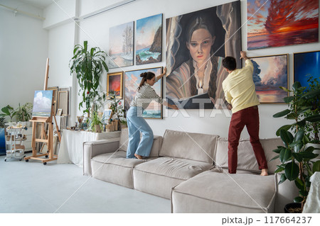 Creative young family couple hangs finished big modern painting on wall in home art workshop. Creative young family couple hangs finished big modern painting on wall in home art workshop. 117664237