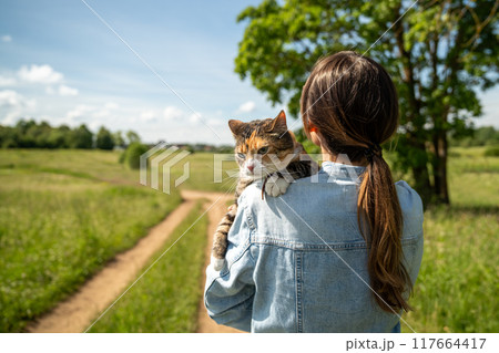 Rear view of woman hugging calm peaceful cat tightly while walking in rural countryside nature area Rear view of woman hugging calm peaceful cat tightly while walking in rural countryside nature area 117664417