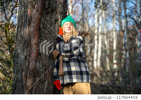 Smiling and relaxed woman strolls in autumn cold forest, warms up in sunny rays and hugs tree trunk Smiling and relaxed woman strolls in autumn cold forest, warms up in sunny rays and hugs tree trunk 117664458