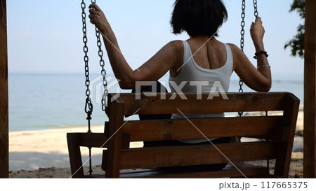 Happy woman ride on swing at tropical at sea beach. Carefree lady enjoying rest near ocean. Adult female having fun at sea shore. Concept of summer vacation or holiday. Close up Slow mo 117665375