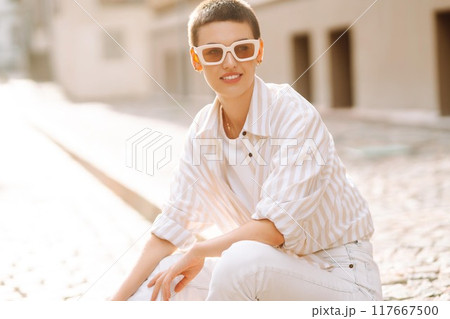 Hipster fashion woman in stylish glasses sits on a curb in the street in Europe. 117667500