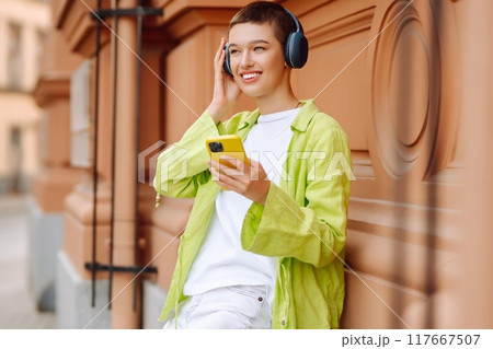 Summer portrait of a young woman with a short haircut with a phone and wireless headphones. 117667507