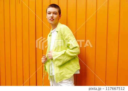 Woman in stylish clothes and short hair poses against orange wall. Fashionable outfit for the city. 117667512