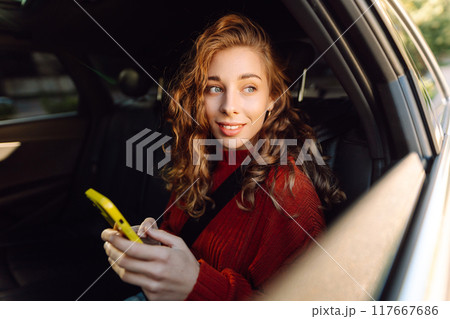 Positive young woman in casual clothes sits in the back seat of a car with a mobile phone. 117667686