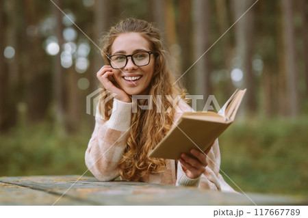 Happy woman is sitting at table with book in beautiful forest. Concept of relaxation, lifestyle. 117667789